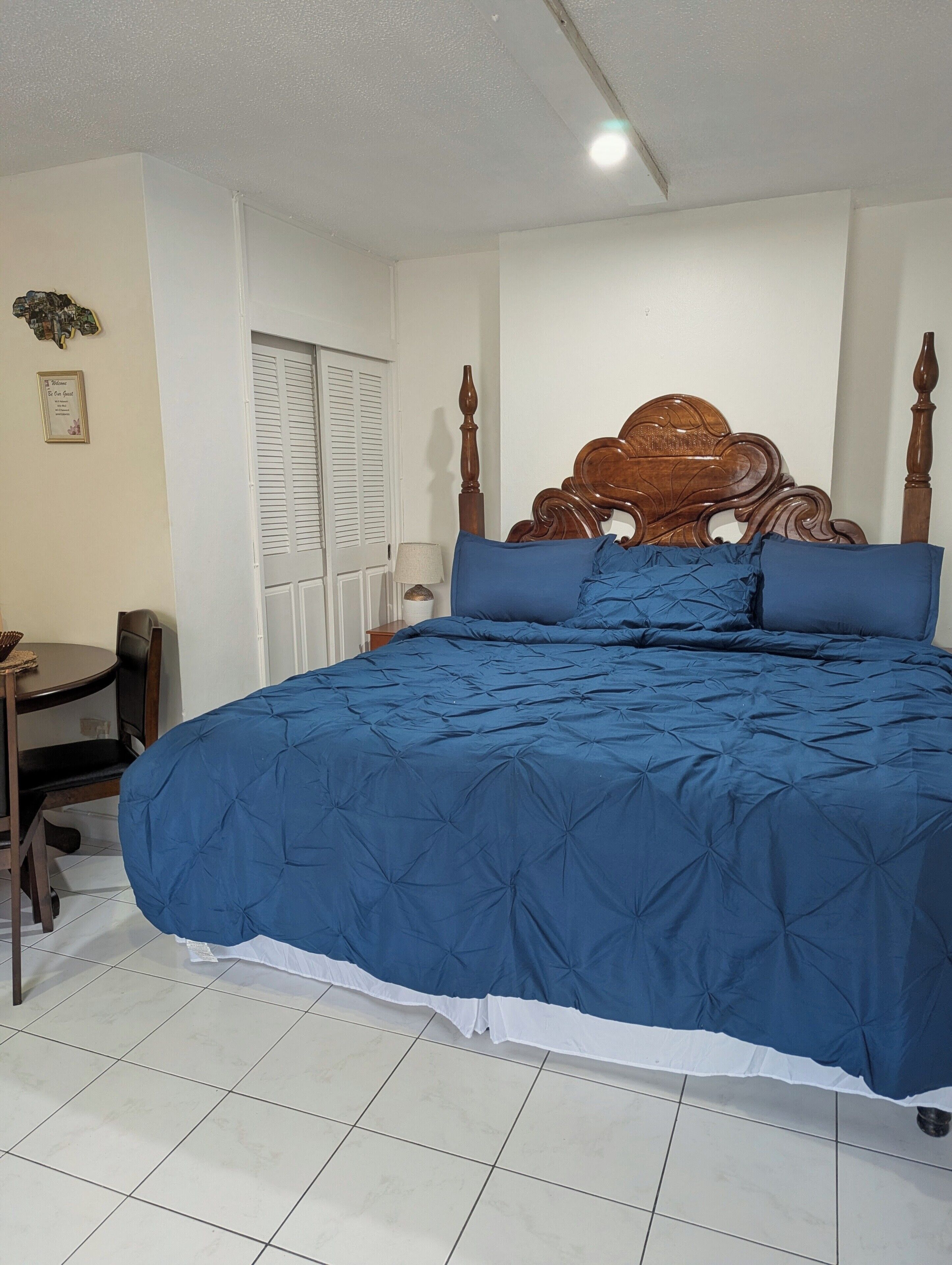 A bedroom with a large bed, a wooden headboard, a bedside table, a chair, and a window with shutters.
