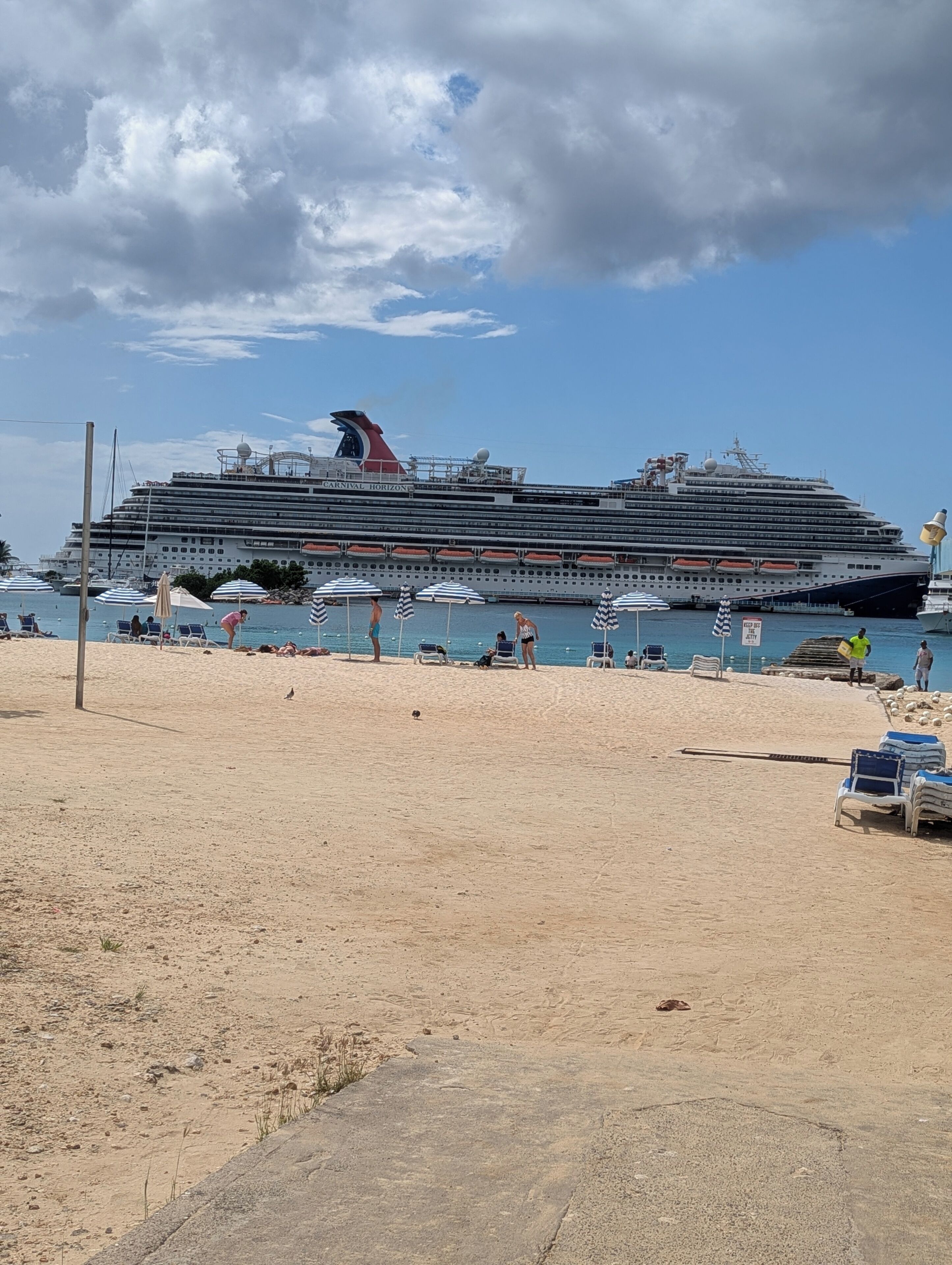 Ein großes Kreuzfahrtschiff liegt an einem Strand mit Liegestühlen und Sonnenschirmen.