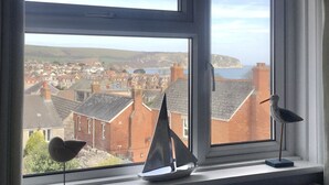 Blick aus einem Fenster auf eine Küstenstadt mit einem Segelbootmodell und zwei vogelartigen Skulpturen auf dem Fensterbrett.