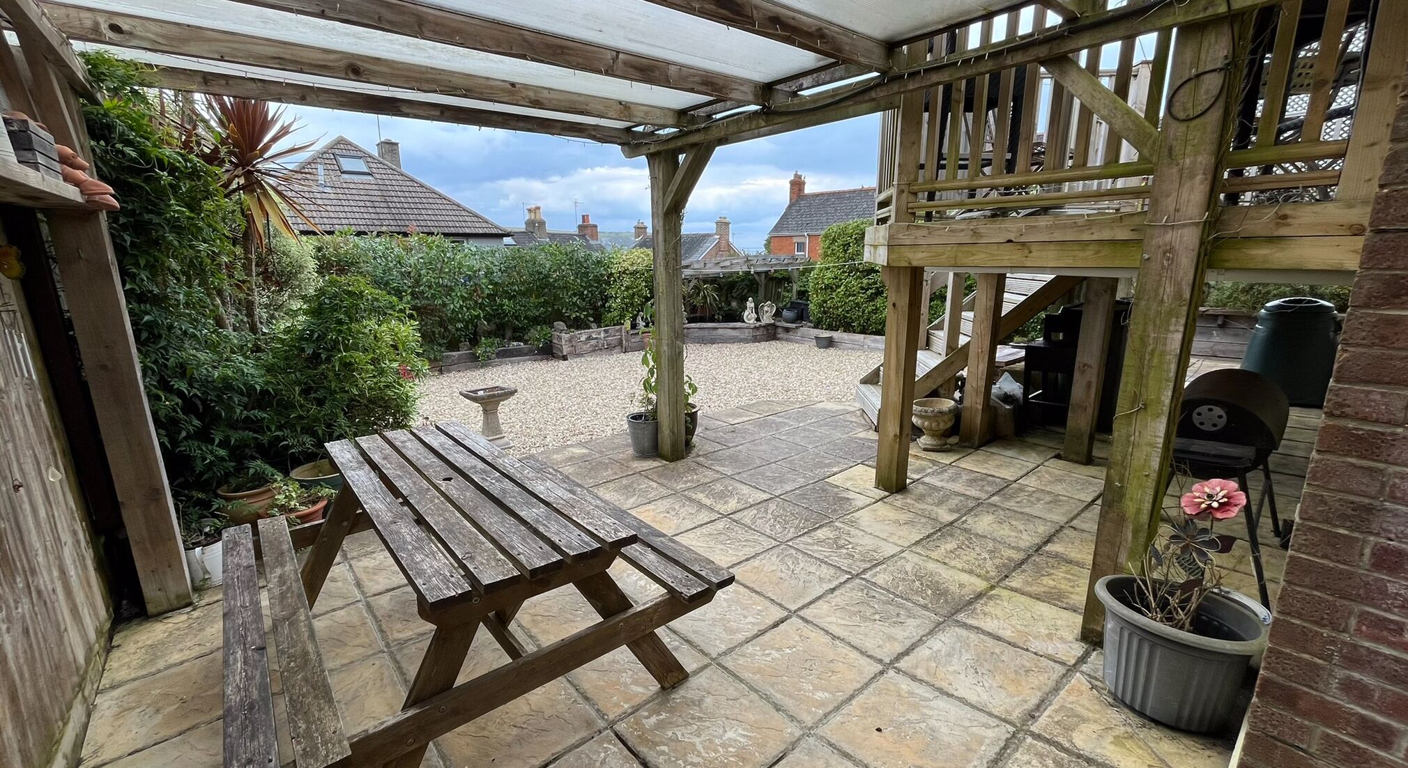 A wooden pergola with a table and chairs, surrounded by potted plants and a gravel area.