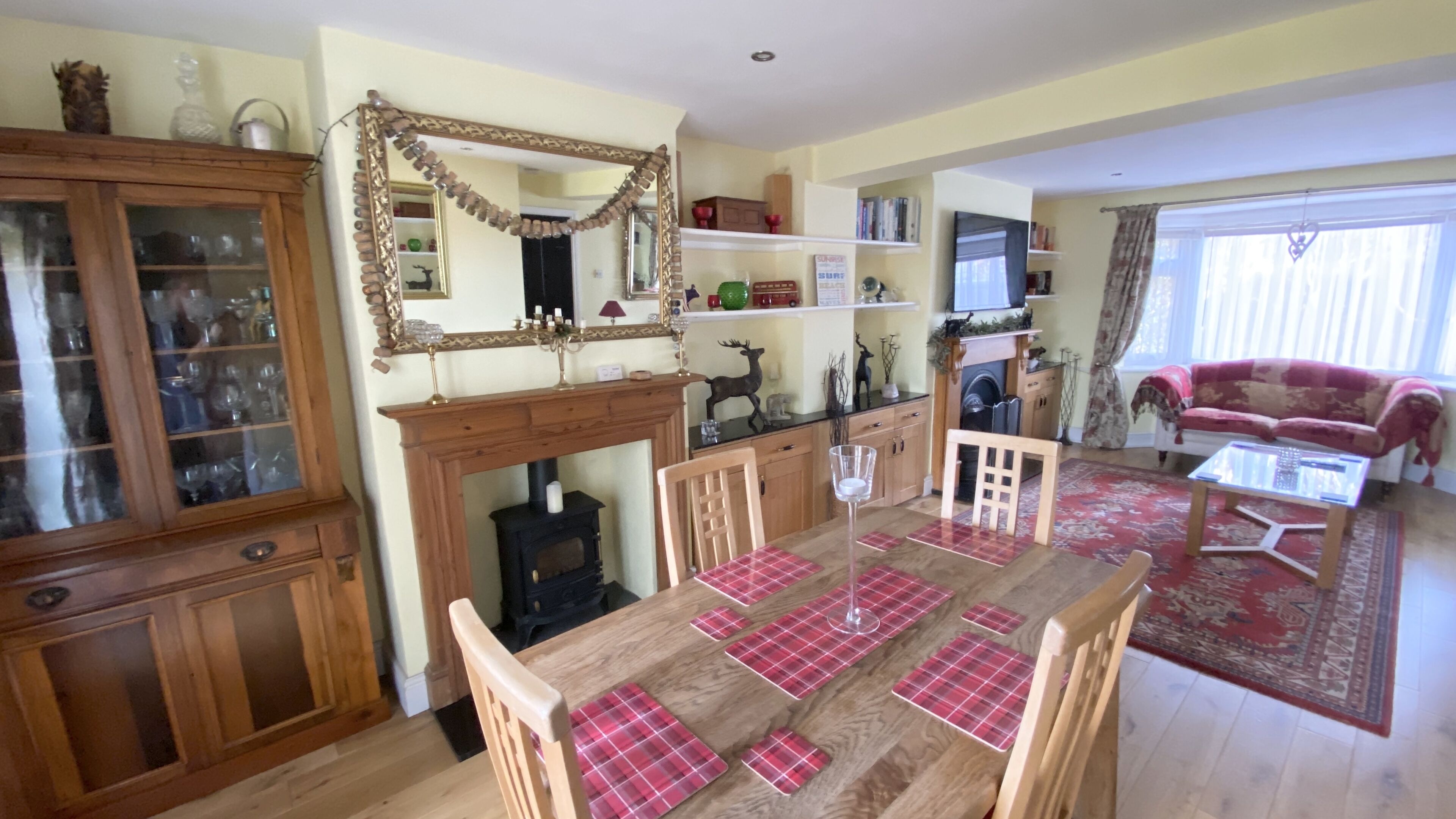 A dining area with a wooden table, chairs, and a red plaid table runner. There is a fireplace with a mantel, a mirror, and shelves with various items. A red sofa and a patterned rug are visible in the background.