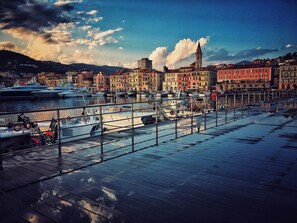 A waterfront promenade with a row of colorful buildings, a prominent tower, and boats docked along the pier.