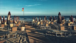 Una playa con filas de tumbonas y sombrillas, un asta con banderas y el océano al fondo.