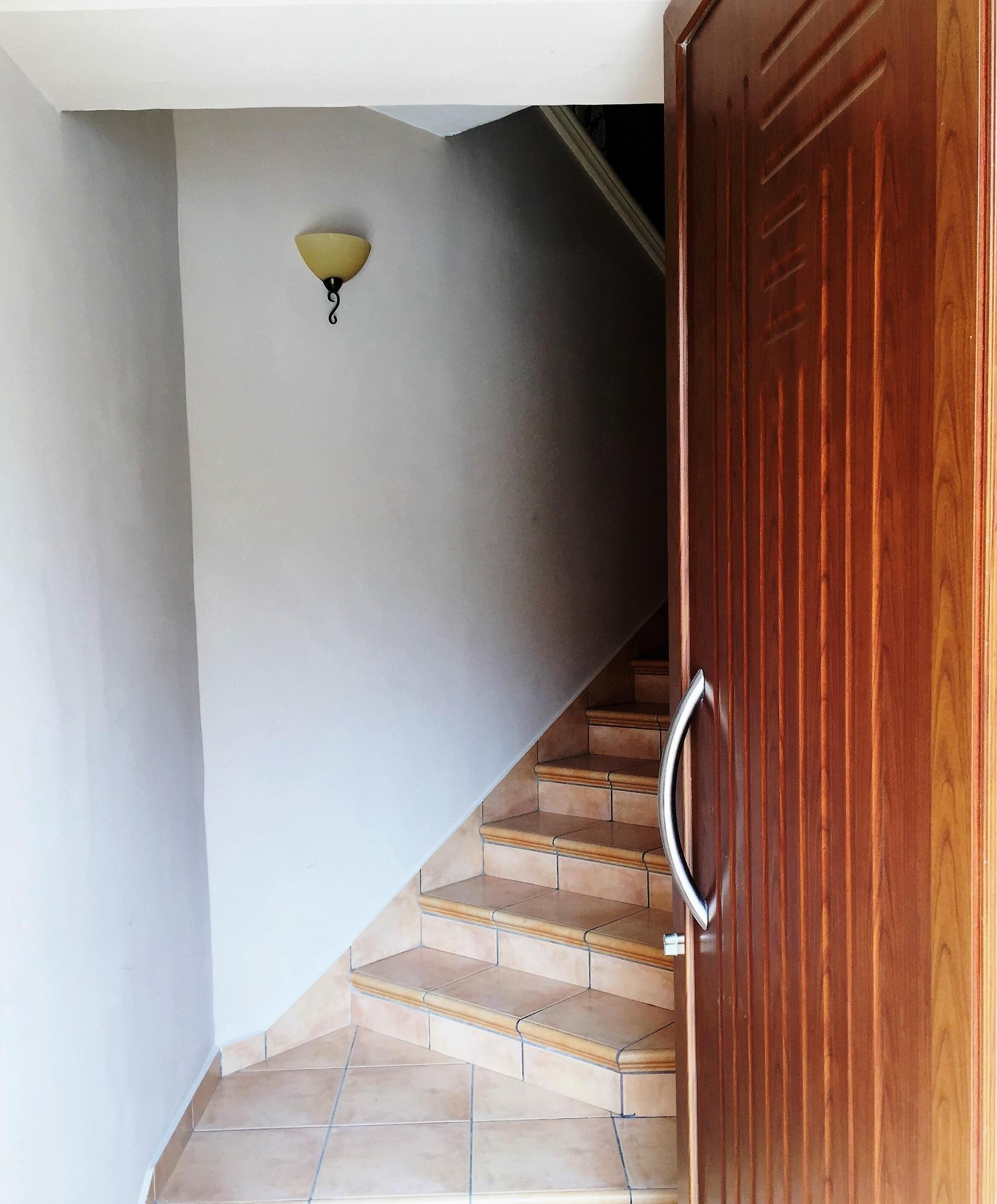 Una escalera de madera con barandilla que conduce a una habitación con puerta de madera y una lámpara fijada a la pared.