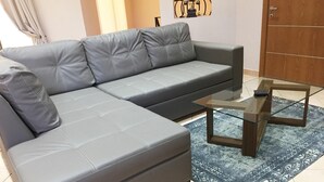 A grey sectional sofa with a matching pillow, a glass-top coffee table, and a patterned rug on a tiled floor.