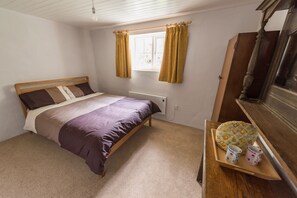 Ein Schlafzimmer mit einem hölzernen Bett, einem Fenster mit Vorhängen, einem Heizkörper und einer Holzkommode mit Spiegel.