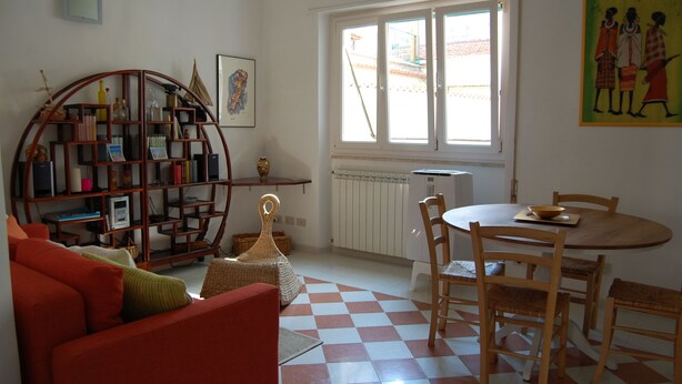 Een woonkamer met een rode bank, een houten boekenkast, een eettafel met stoelen en een geruit vloerkleed.