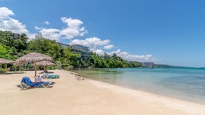 A beach with lounge chairs and a thatched umbrella, a clear blue sky, and buildings in the background. -  Waterfront ,  minutes from the beach, self catering, 24 hr security,            (Ocho Rios)