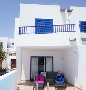 A two-story building with blue shutters, a balcony with a metal railing, and a seating area with a grey sofa and two chairs.
