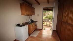 A compact kitchen with wooden cabinets, a white refrigerator, and a coffee maker. A small dining area with a table and chairs is visible through an open door. - Quinta do Pântano is a calm and reserved place, with unique ambience and landscapes (Machico)