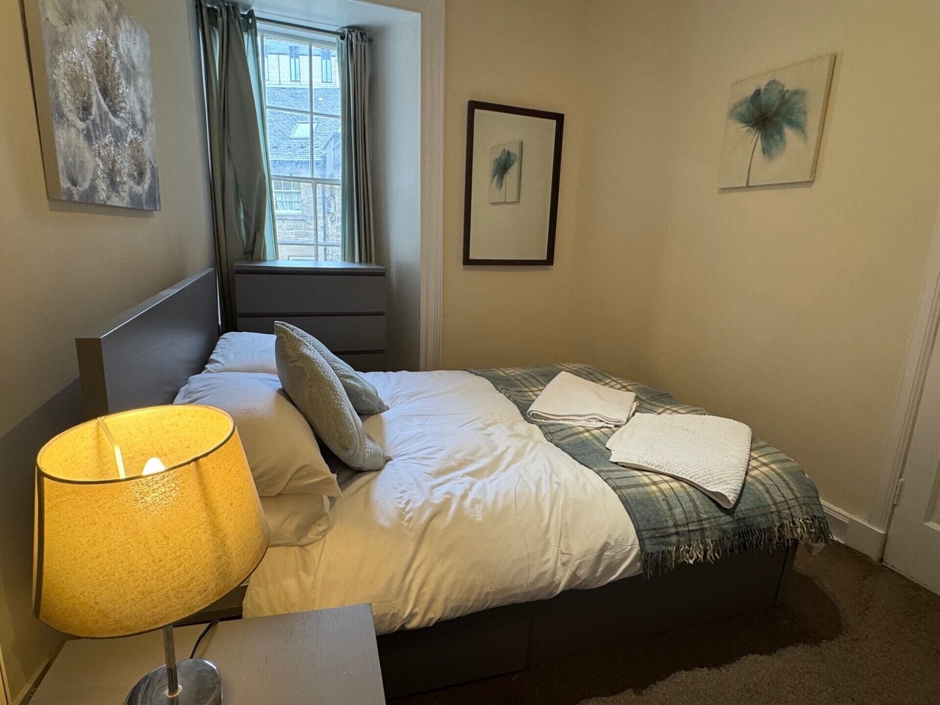 A neatly made bed with a plaid blanket, a bedside lamp, and a window with curtains.