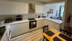 Eine moderne Küche mit weißen Schränken, einem Einbauofen, einer Mikrowelle und einem Toaster. Ein Esstisch mit Stühlen und ein an der Wand montierter Feuerlöscher sind ebenfalls vorhanden.