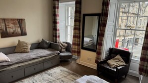 Ein gemütliches Zimmer mit einer Couch, einem Sessel, einem Bett, einem großen Spiegel und Blick auf die Stadt durch ein Fenster.