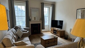 Ein Wohnzimmer mit einer Sofagarnitur, einem Fernseher, einem Kamin und einem großen Fenster mit Vorhängen.