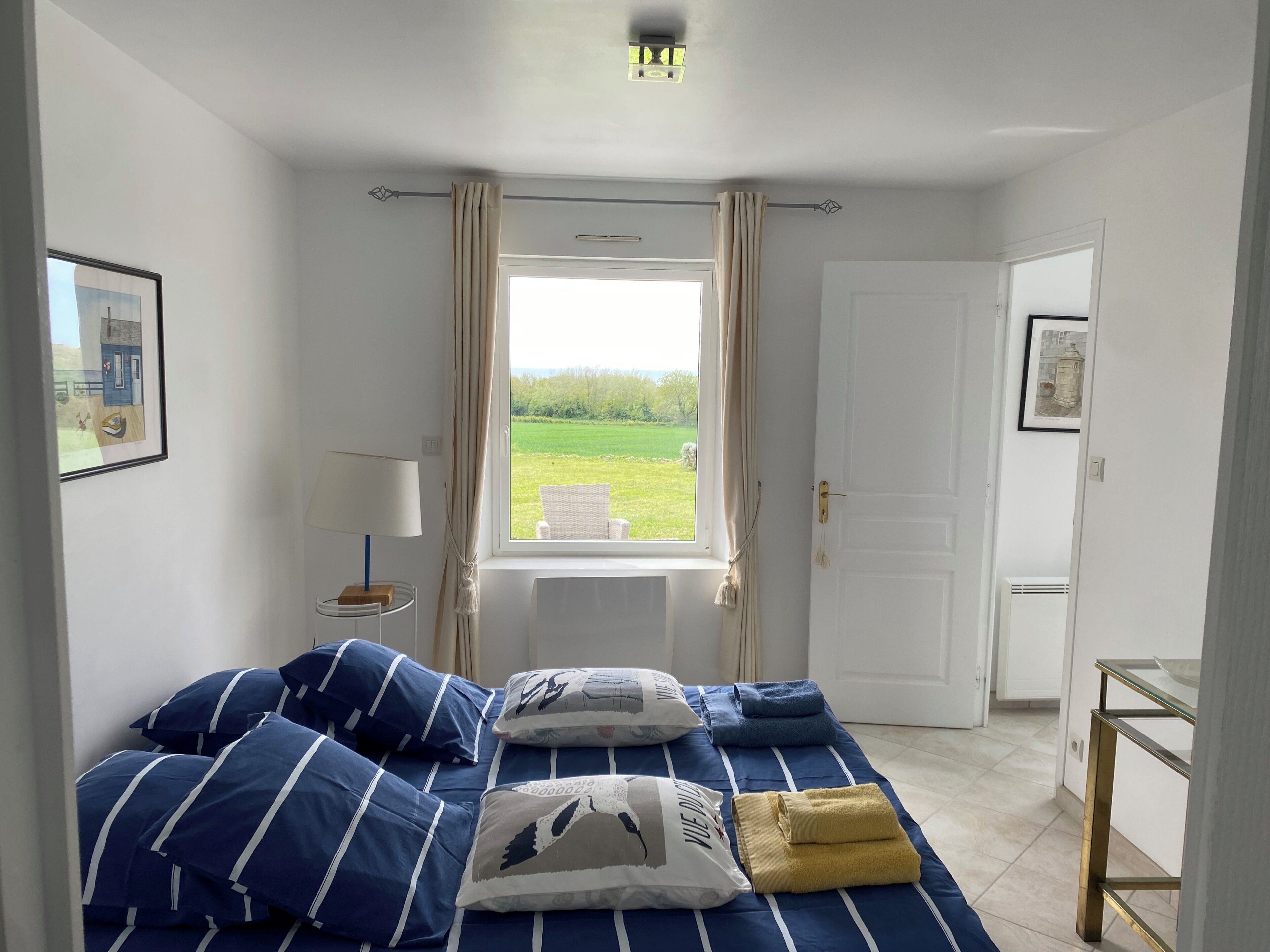 Une chambre à coucher avec un lit, une fenêtre avec des rideaux, une table de chevet, une lampe et un tableau encadré au mur.