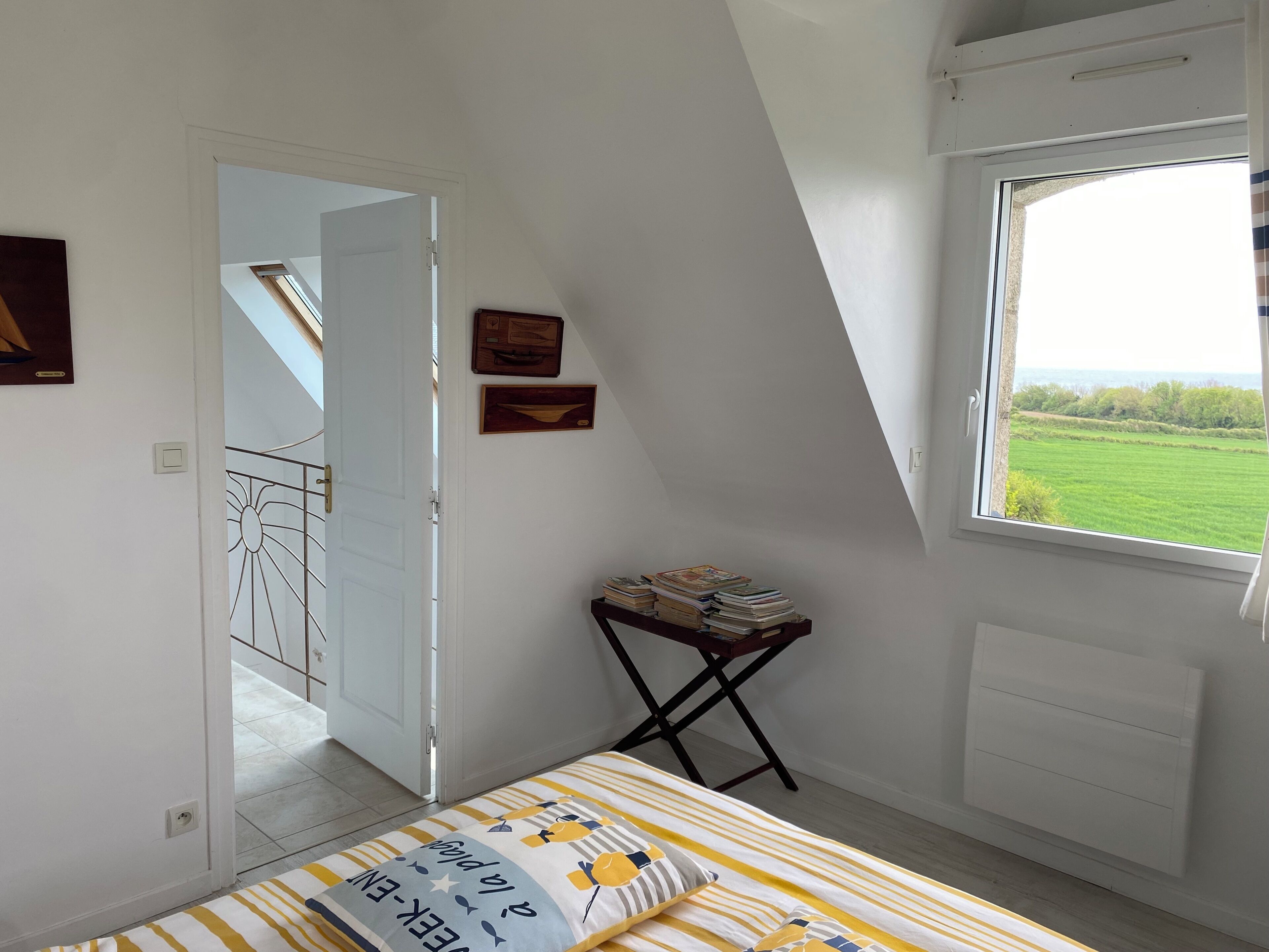 A bedroom with a bed, a window with a view of a green field, a door leading to another room, and a small table with books.