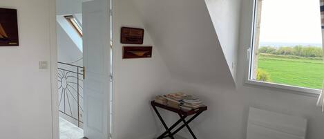 A bedroom with a bed, a window with a view of a green field, a door leading to another room, and a small table with books.