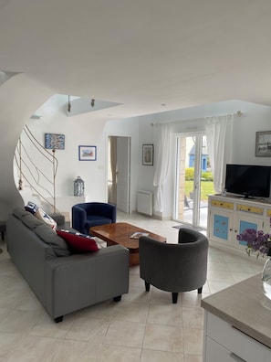 A modern living room with a grey sofa, blue armchair, and a coffee table. There is a TV on a cabinet, and a staircase leading to another level. - 180° SEA VIEW, BEACH NEARBY FOR THIS SPACIOUS ARCHITECT-DESIGNED HOUSE (Primelin)
