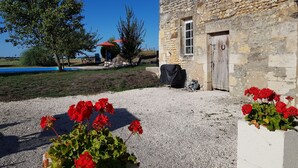 A stone building with a door, a gravel area with red flowers, and a pool in the background.
