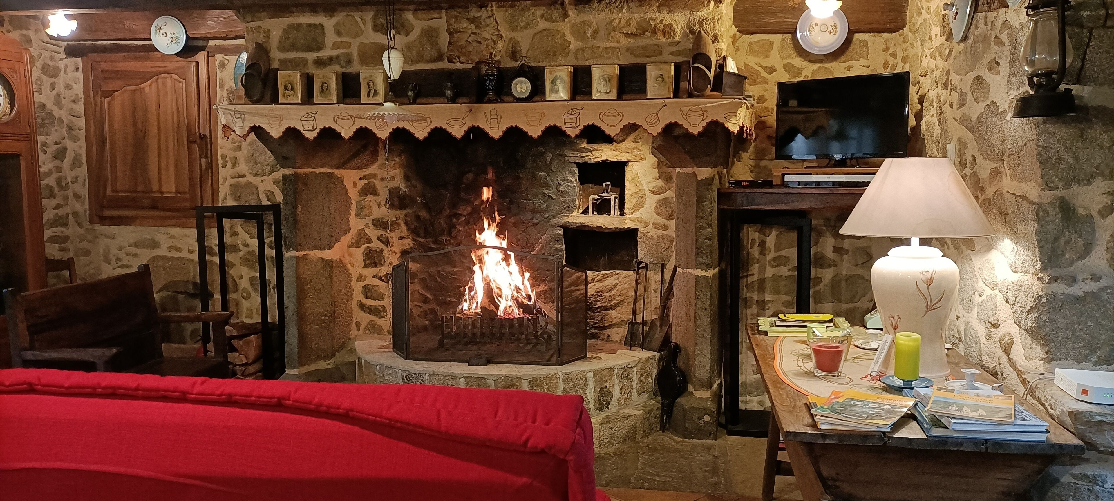 A stone fireplace with a fire, wooden furniture, and a table with books and a lamp.