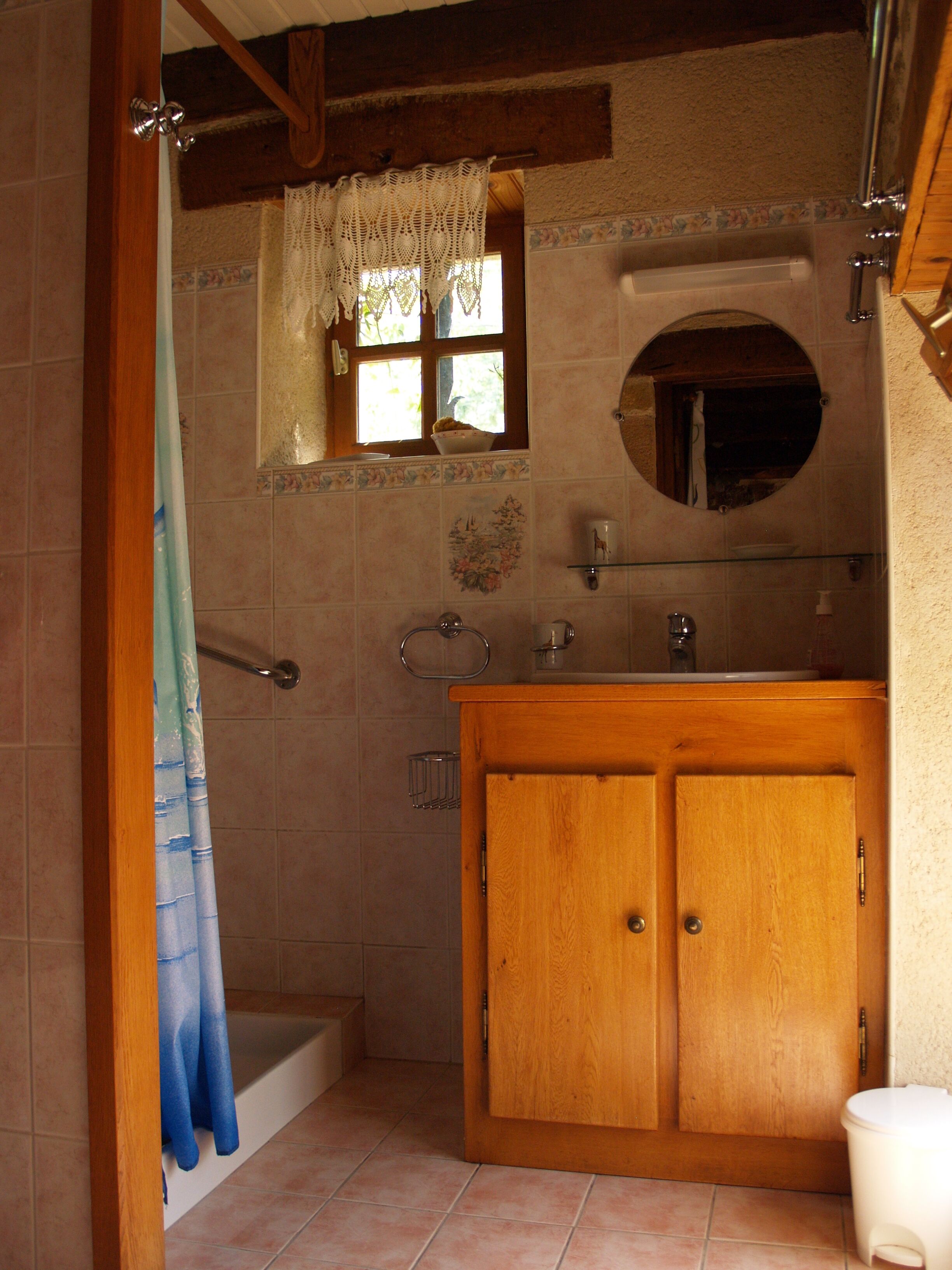A bathroom with a wooden vanity, a small window with a crocheted curtain, a round mirror, and a bathtub.