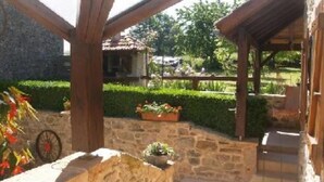 A stone patio with a wooden pergola, surrounded by well-maintained hedges and a view of a distant building. - country holiday home - LABASTIDE L'EVEQUE (Le Bas Ségala)