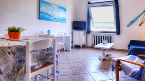 Et lille, godt oplyst rum med flisegulv, et spisebord med stole, en sofa, et fjernsyn på et skab, et vindue med gardiner og et billede på væggen.