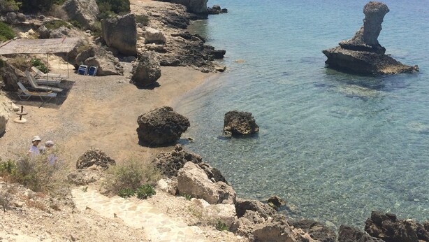 Ein felsiger Strand mit klarem Wasser, einigen Liegestühlen und einem Felsen im Meer.