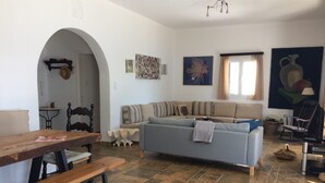 Ein Wohnzimmer mit einer Couch, einem Holztisch und einer Bank. Ein Fenster mit Vorhängen, ein Wandregal mit einem Hut und ein Gemälde an der Wand sind ebenfalls zu sehen.