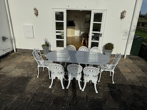 Une terrasse avec une table ronde et des chaises blanches, une porte blanche et deux plantes en pot.