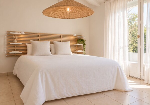 A bedroom with a large bed, a hanging light fixture, a bedside table with a plant, and a view of greenery outside.