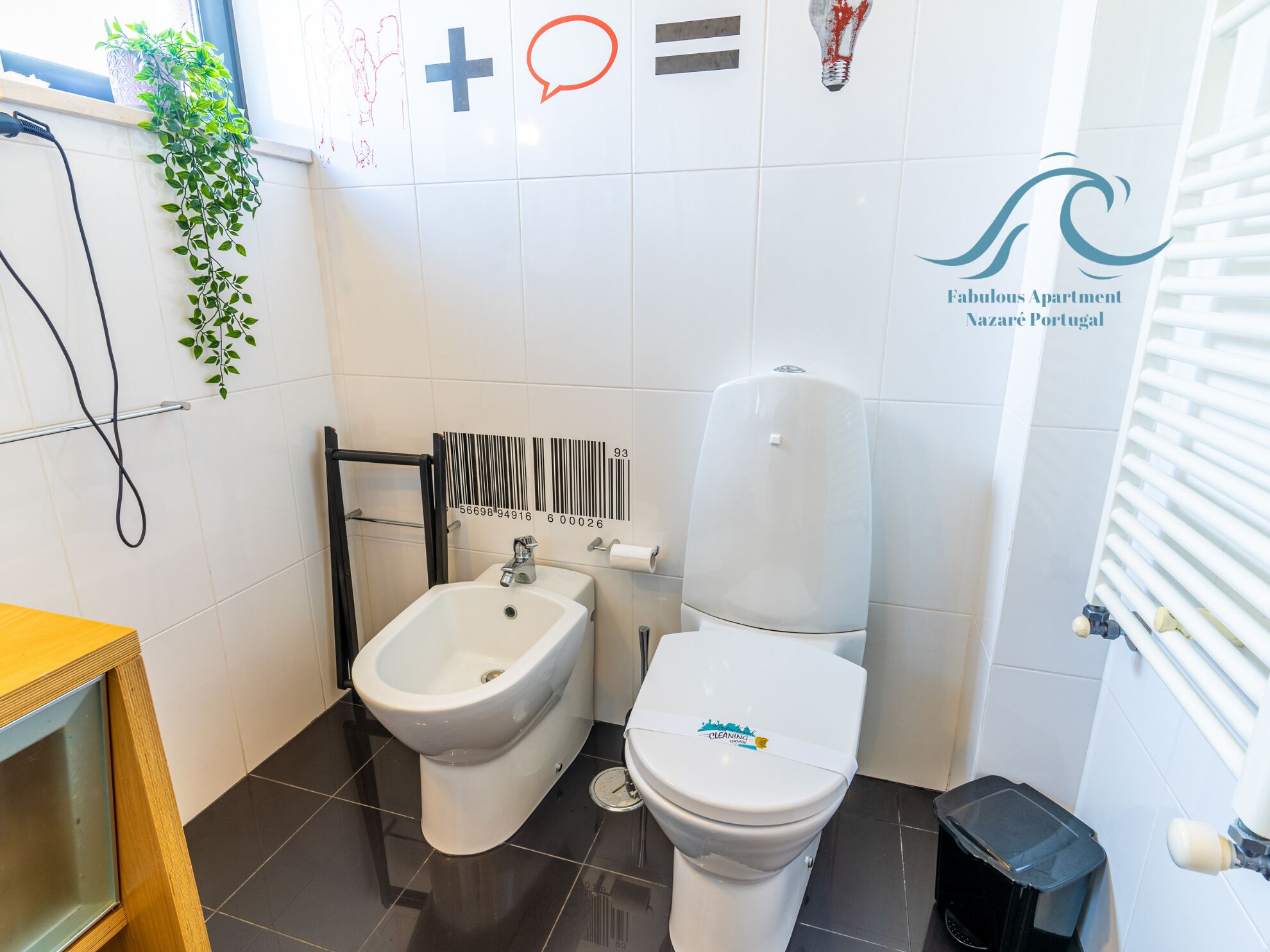 A bathroom with a bidet, toilet, and a hand dryer. There is a plant on the wall and a sign with a logo.