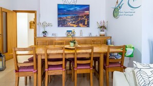 Een eetruimte met een houten tafel en stoelen, een hanglamp, een stadsgezicht schilderij en een klok aan de muur.