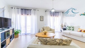 Een moderne woonkamer met een bank, een salontafel en een televisie. Gordijnen, een kroonluchter en een logo op de muur.