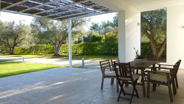 An outdoor patio with a wooden table and chairs, surrounded by a garden with trees and a paved walkway.