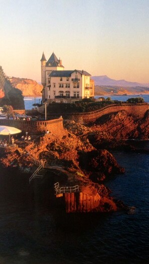 A castle-like building with a prominent tower, situated on a rocky coastline with a body of water in the foreground.