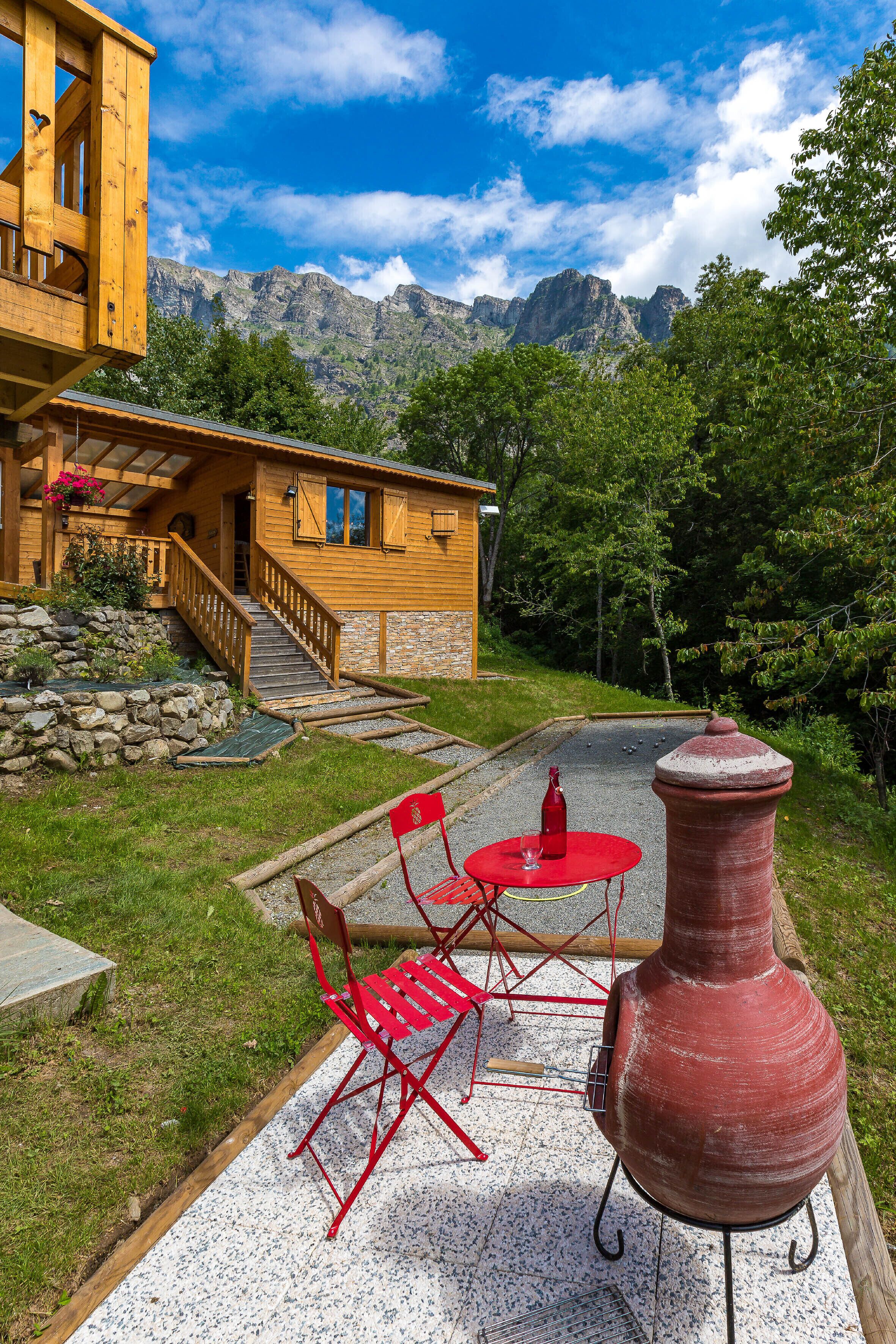 Una cabaña de madera con una escalera de piedra, una mesa y silla rojas en el exterior, y una maceta de cerámica grande.