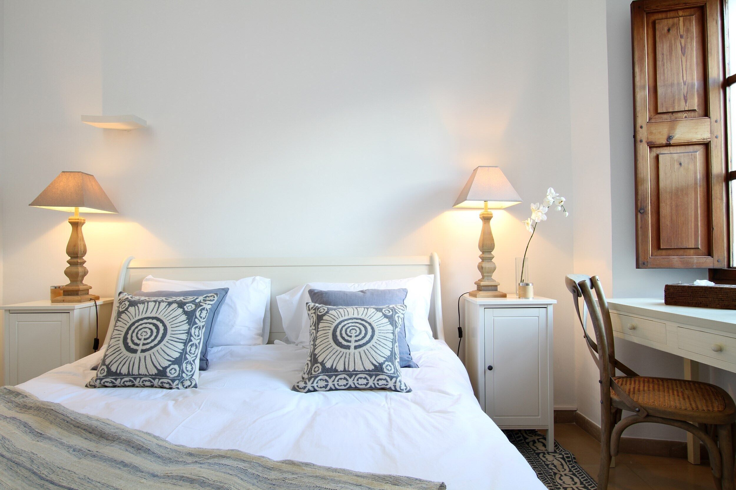 Een net opgemaakt bed met twee decoratieve kussens, een nachtkastje met een lamp, een houten stoel en een bureau met een lamp.