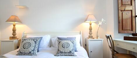 Een net opgemaakt bed met twee decoratieve kussens, een nachtkastje met een lamp, een houten stoel en een bureau met een lamp.