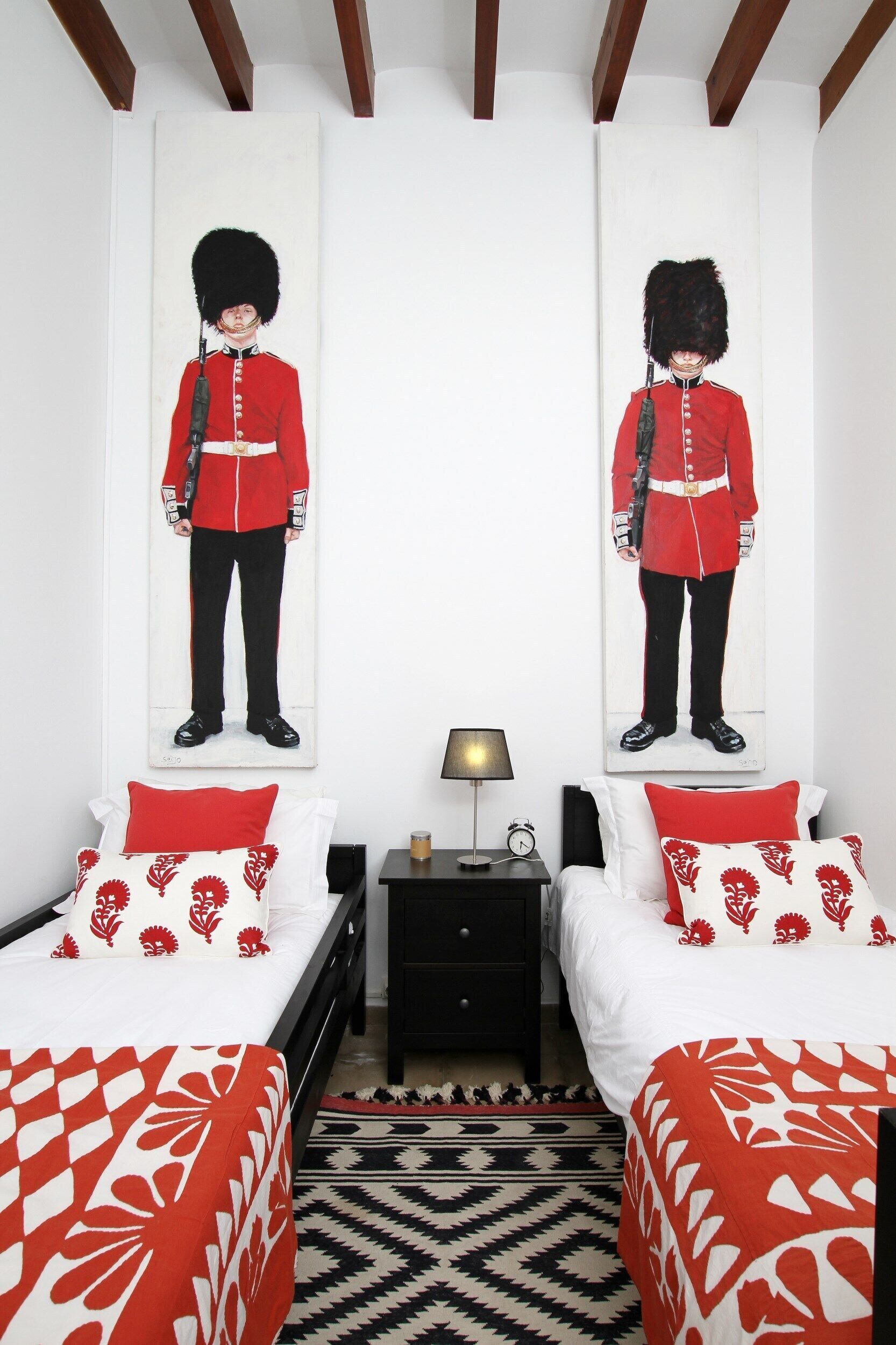 A bedroom with two beds, each with red and white bedding, a black and white patterned rug, and a nightstand with a lamp between the beds.