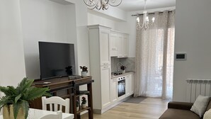Un salon moderne avec un canapé marron, une table à manger blanche et des chaises. Un téléviseur à écran plat est fixé au mur, et il y a une cuisine avec des armoires blanches et une cuisinière.