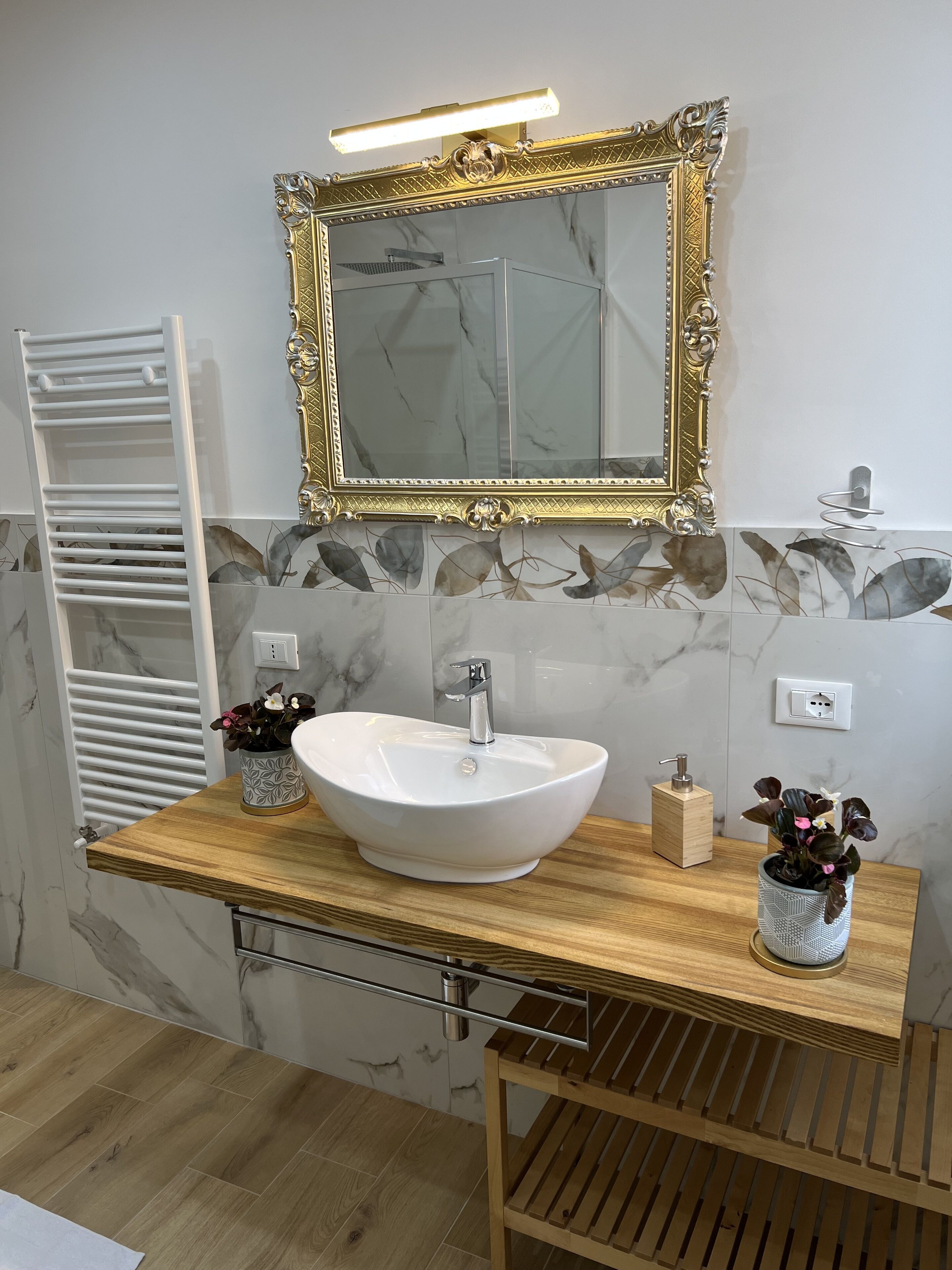 Ein Badezimmer mit einem weißen Waschtisch, einem spiegelnden Spiegel mit goldener Rahmen, einer Holzkommode, einem Handtuchhalter und einem Topf mit Pflanze.