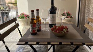 Une table de balcon avec une bouteille de vin, deux verres à vin, un saladier de fraises et deux plantes en pot aux fleurs roses.