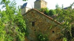 Un bâtiment en pierre, surmonté d’un toit de tuiles, entouré de verdure et baigné d’un ciel dégagé.