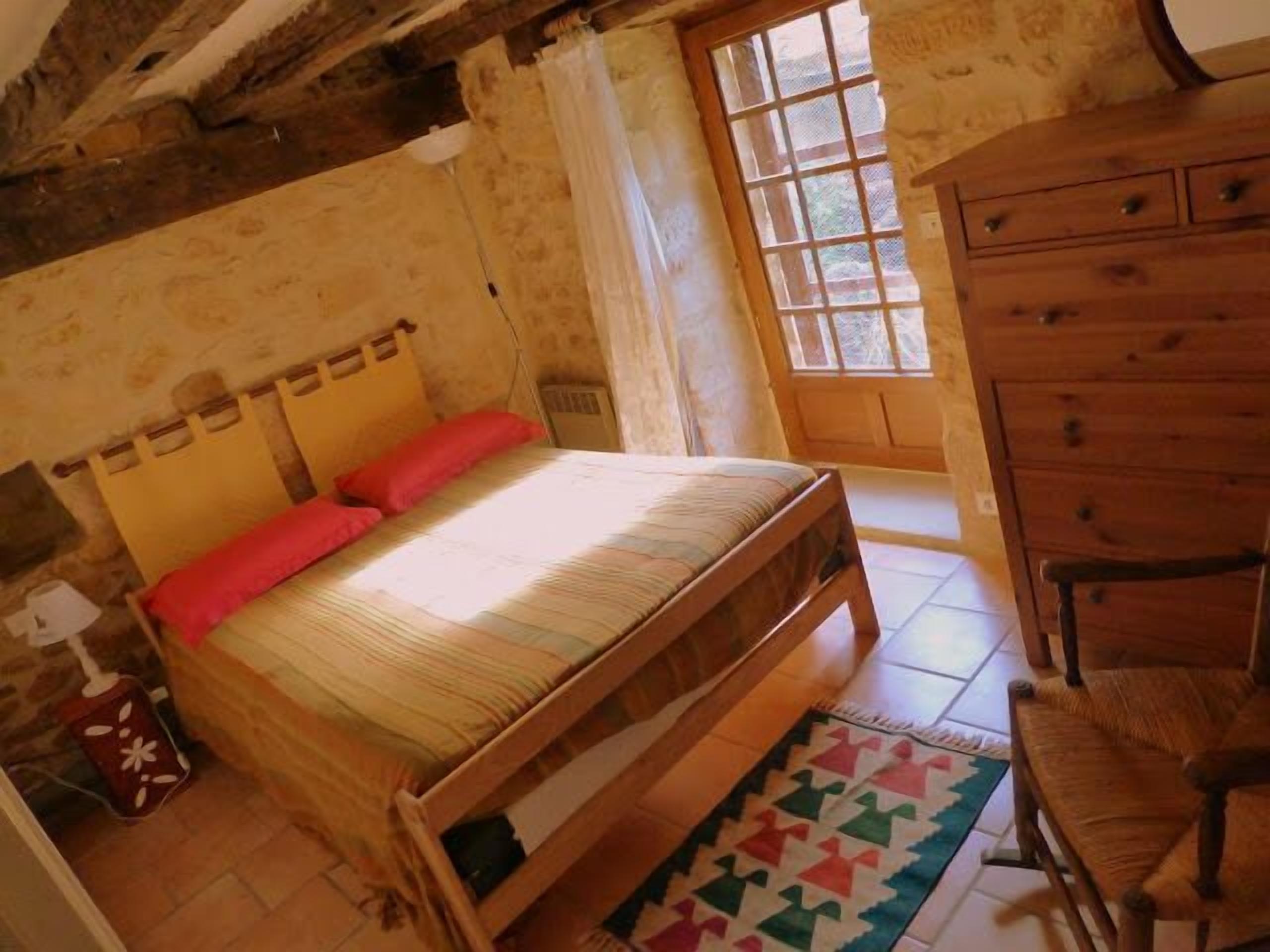 Een houten bed met hoofdeinde, een houten dressoir en een stoel in een kamer met een betegelde vloer en een raam met uitzicht.