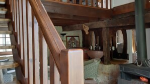 Un intérieur rustique avec un escalier en bois, un foyer en pierre doté d’un poêle à bois, et divers ustensiles de cuisine.