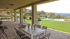 Eine Außenpatio mit Steinboden, einem weißen Tisch und Stühlen. Es gibt Kübelpflanzen und einen Blick auf ein Schwimmbad und die Landschaft.
