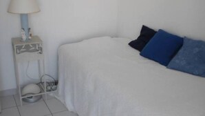 A small, neatly arranged bedroom with a bed, a bedside table with a lamp, and a small shelf with decorative items.