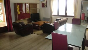 Ein Wohnzimmer mit einer Couch, Sesseln, einem Glastisch, einem Fernseher und einem Schrank.