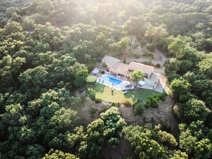 Vue aérienne d’une maison entourée d’une forêt dense, avec une piscine et un espace de détente extérieur.